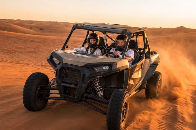 Couple in helmets self-driving a 2-seater dune buggy across Dubai red dunes at sunset