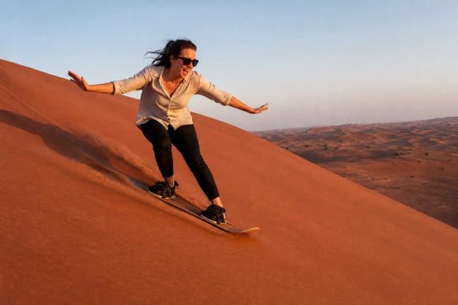Guest sandboarding down a red Dubai desert dune, arms out and laughing