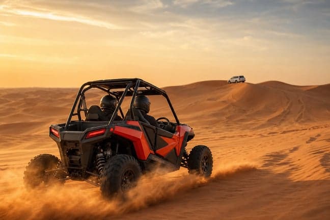 Polaris RZR dune buggy mid-drive on red sand dune with dust trail and Land Cruiser in distance