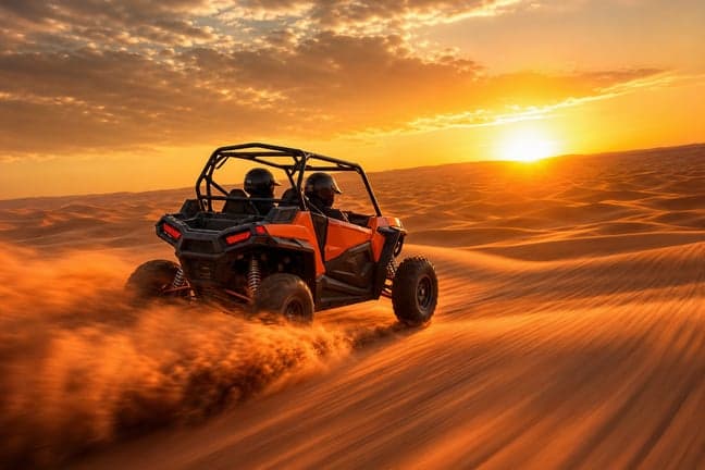 Orange Polaris RZR 2-seater dune buggy with helmeted couple driving on red sand dune at golden hour