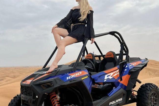 Blonde woman sitting on blue and orange Polaris RZR XP 1000 buggy roll cage in Dubai desert