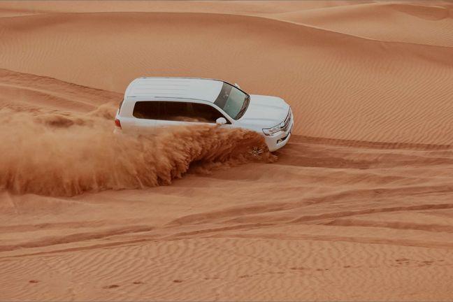 White Toyota Land Cruiser descending steep red sand dune with spray behind in aerial view of Dubai desert