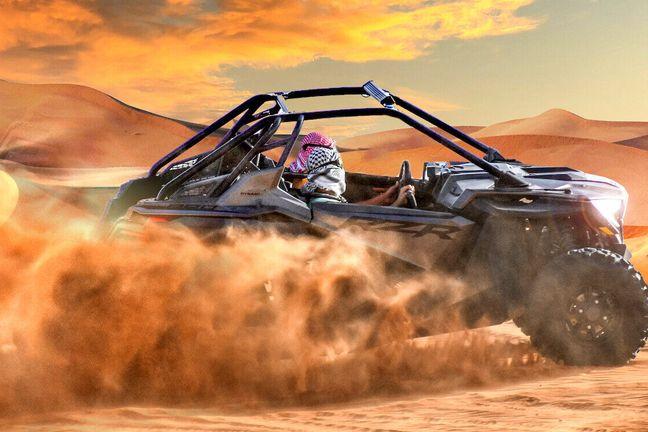 Driver wearing traditional keffiyeh headscarf driving Polaris RZR buggy through orange sand spray at sunset