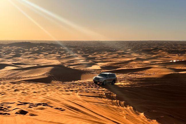 Gray Land Cruiser navigating vast golden Dubai desert dunes at sunset with sun rays and lens flare
